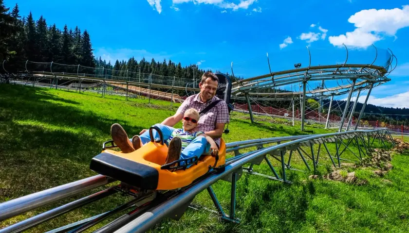 Ojciec i syn z uśmiechem zjeżdżają na sankach po torze zjazdowym. W tle widać czarną górną część konstrukcji toru.