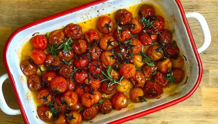 Tomates cherry confitados en aceite de oliva, con romero y tomillo, en una bandeja blanca con borde rojo.