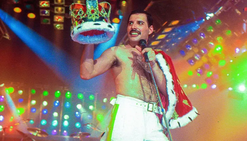 Freddie Mercury, w koronie i z mikrofonem, na scenie pełnej świateł. Pamięć o nim żyje, choć zmarł Freddie Mercury.