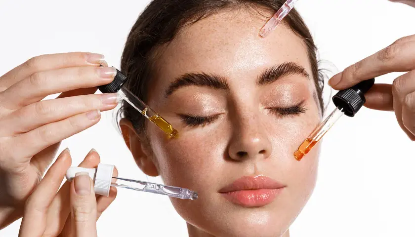 Manos aplican sueros con goteros en el rostro de una mujer. El brillo en su piel sugiere el poder de la vitamina C para la cara.