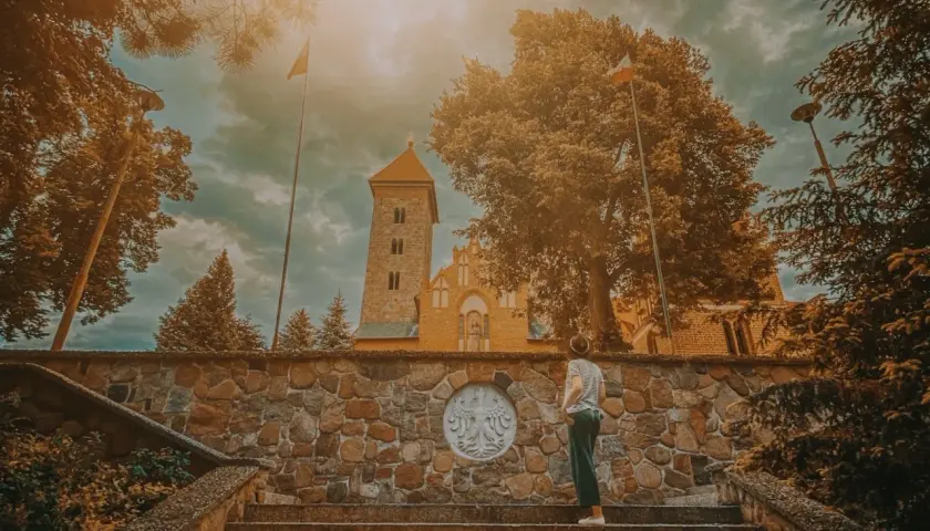 Osoba na schodach przed kamiennym murem z godłem Polski, w tle kościół w Czerwińsku nad Wisłą.