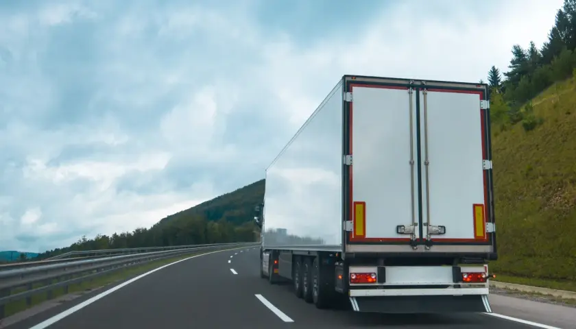 Ciężarówka na autostradzie, z tyłu widać białą naczepę. Na poboczu rosną drzewa, a nad nimi chmury. Tablice wyróżniające przepisy są widoczne na naczepie.