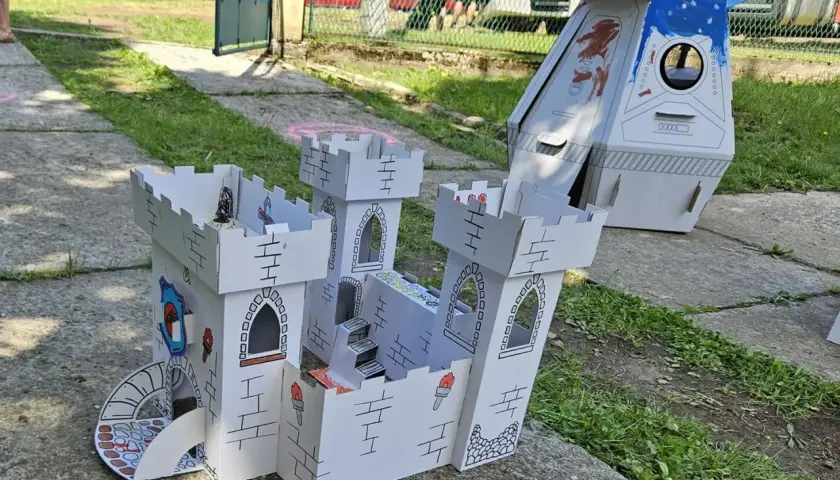 Zabawki z kartonu diy: biały zamek z wieżami i schodami, obok rakieta. Dzieci bawią się w tle.
