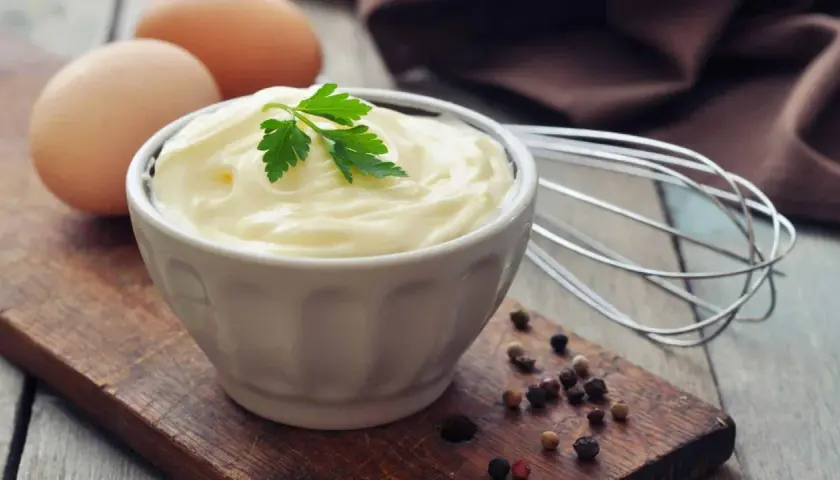 Frische Eier und Petersilie auf einem Holzbrett, bereit, um köstliche Mayonnaise selber zu machen. Ein Schneebesen liegt daneben.