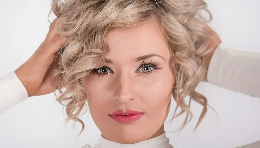 Kobieta z kręconymi blond włosami, która nonszalancko układa je dłońmi. Jej spojrzenie jest hipnotyzujące, a usta pomalowane na różowo.