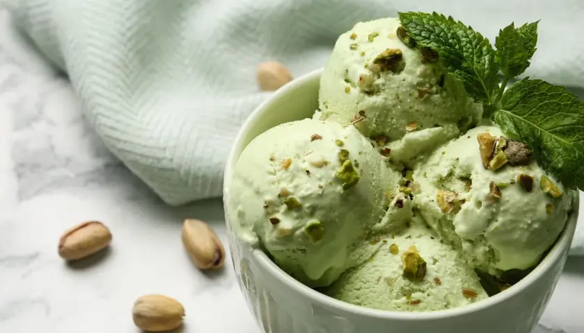 Coppa di cremoso gelato al pistacchio, guarnita con foglioline di menta e granella di pistacchi. Alcuni pistacchi interi sparsi sul marmo.