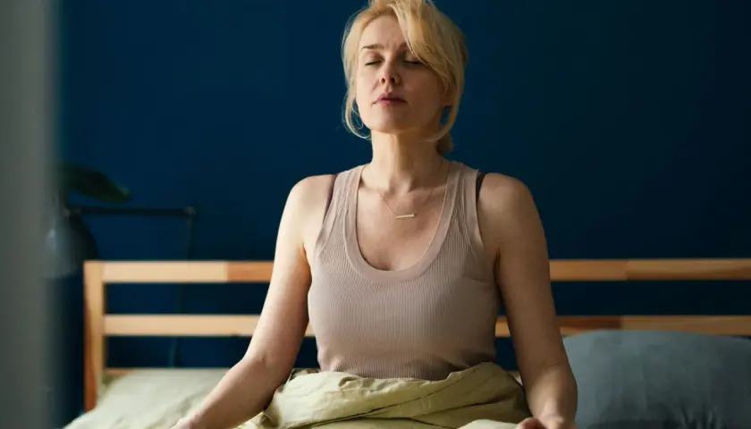 Kvinna med slutna ögon sitter i sängen, insvept i täcke, i en avslappnad somna meditation.