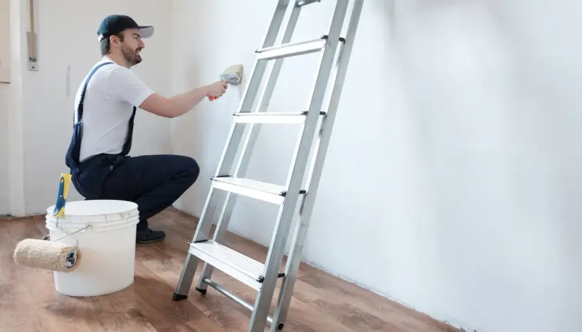 Hombre con brocha y cubo de pintura, listo para limpiar pared antes de pintar. Escalera metálica al lado.