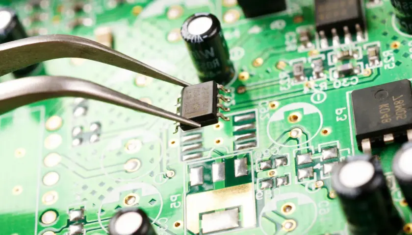 Pęseta trzyma układ scalony na zielonej płytce drukowanej. Widać elementy elektroniczne, kondensatory i ścieżki. Zbliżenie ujawnia potencjalne oznaki korozji elektrochemicznej.