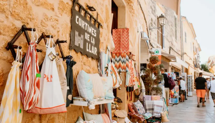 Bunte Stoffe und Kissen hängen vor einem Geschäft am alcudia markt. Menschen schlendern die Gasse entlang.