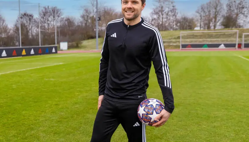 Uśmiechnięty mężczyzna w stroju sportowym trzyma piłkę na boisku. To jego pierwszy staż trenerski, pełen nowych wyzwań i nauki.