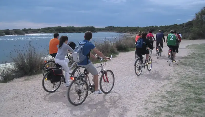 Grupo de ciclistas pedaleando por un camino de tierra junto a la albufera de Valencia. El cielo está nublado y el agua refleja la luz.