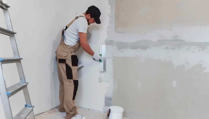 Operaio con tuta beige e cappellino nero mentre impara come intonacare un muro, usando una spatola. Scala e secchio a terra.