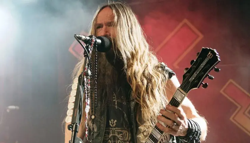 Zakk Wylde, wokalista i gitarzysta, z długimi blond włosami i naszyjnikami, gra na gitarze podczas koncertu.