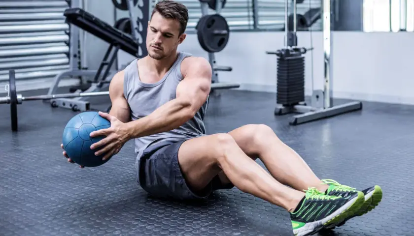 Hombre sentado en el suelo del gimnasio, girando el torso con una pelota medicinal azul, trabajando la elasticidad muscular.