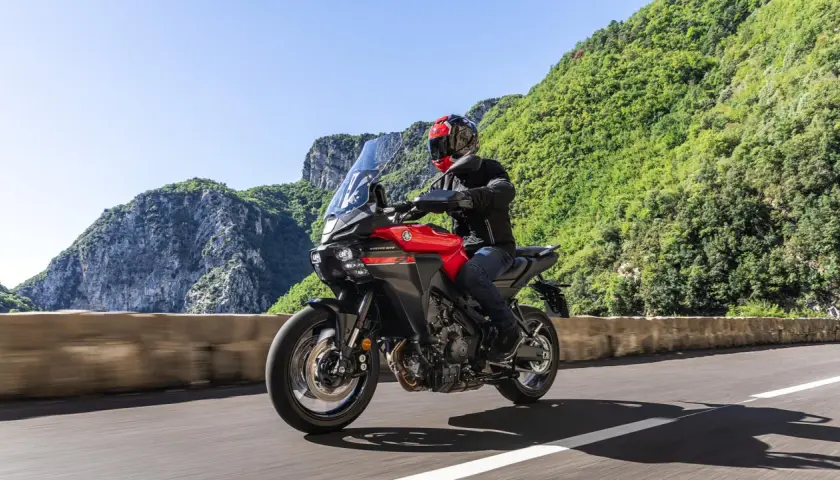 Un motociclista su una Yamaha Tracer rossa affronta una curva su una strada panoramica, godendosi una gita in moto tra montagne verdi e cielo azzurro.