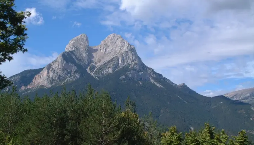 Majestuoso Pedraforca se alza imponente, su altitud desafía las nubes. Bosques verdes cubren sus faldas bajo un cielo azul.