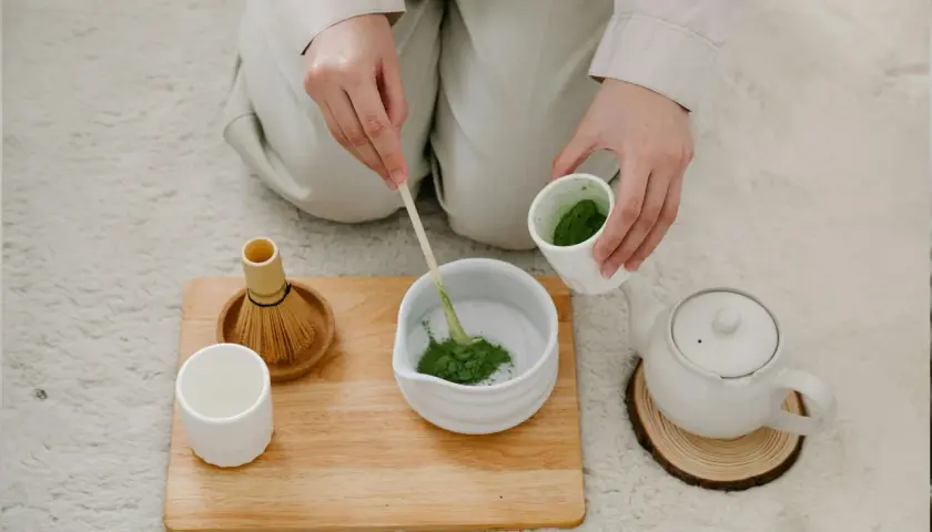 Hände bereiten Matcha zu. Ein Bambusbesen und eine Tasse stehen bereit. Dieser Matcha macht wach!
