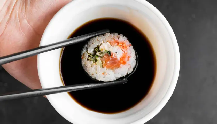 Pałeczkami zanurzam rolkę sushi z łososiem w ciemnym sosie.