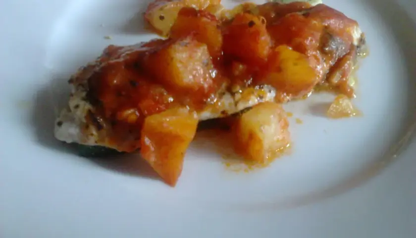 Un delicioso jargo pescado cubierto con salsa de tomate y trozos de patata, servido en un plato blanco.