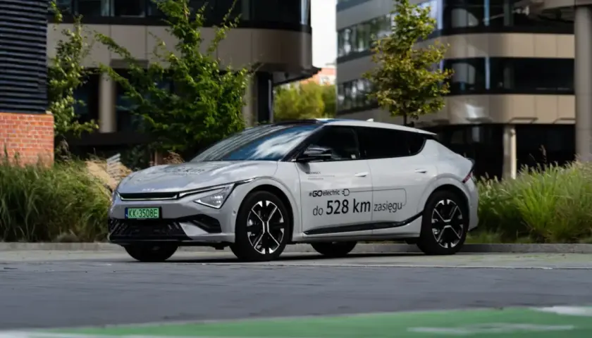 Biały Kia EV6 zaparkowany przed nowoczesnym budynkiem, z napisem "#GOelectric do 528 km zasięgu".