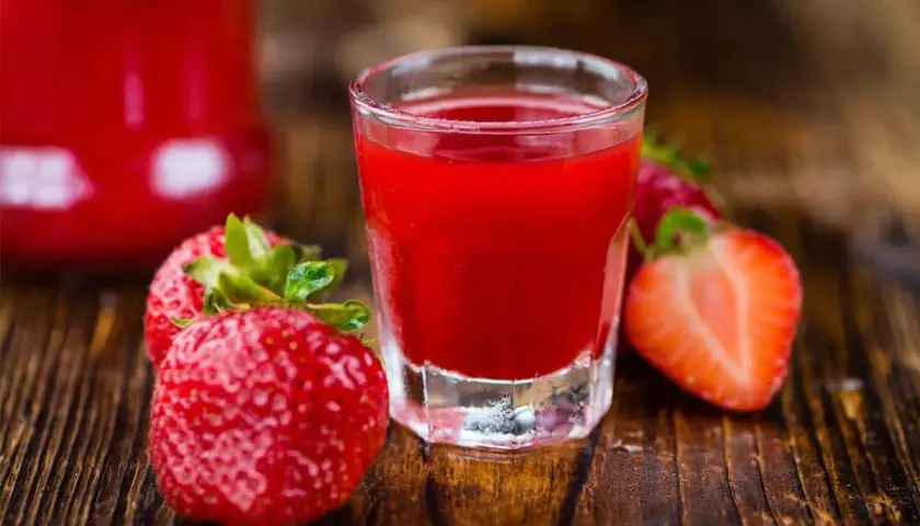 A shot glass filled with vibrant red nalewka z truskawek, surrounded by fresh strawberries on a wooden surface.