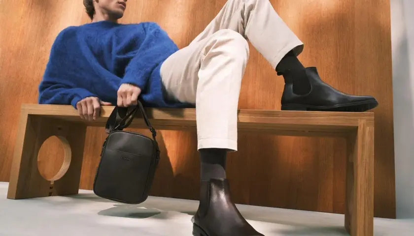 Mężczyzna w niebieskim swetrze i beżowych chinosach prezentuje stylowe buty do chinosów. Elegancka torba na ramię dopełnia całość.