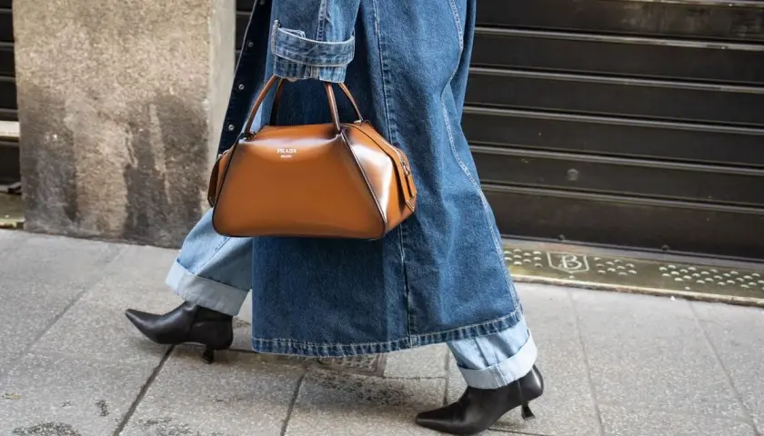 Une femme élégante marche dans la rue, portant un jean retroussé, une veste en jean et un sac à main marron. Le style "comment retrousser son jean femme" est parfait pour cette tenue.