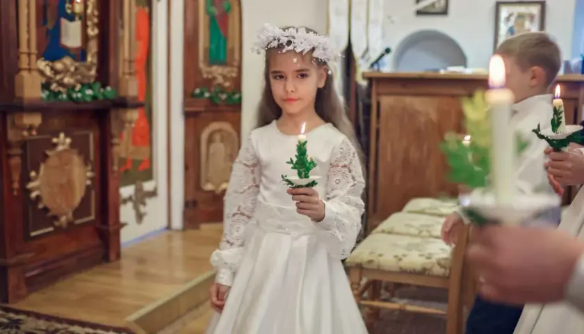 Dziewczynka w białej sukni i wianku trzyma świecę. To ważny dzień, w której klasie komunia.
