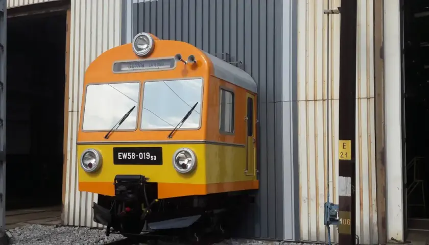 Pomarańczowo-żółty pociąg EW58-019sa stoi przed halą.