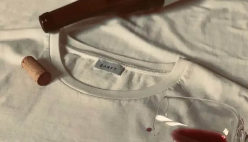 Un bouchon de liège et une bouteille de vin à côté d'un tee shirt blanc, avec une tache de vin rouge.