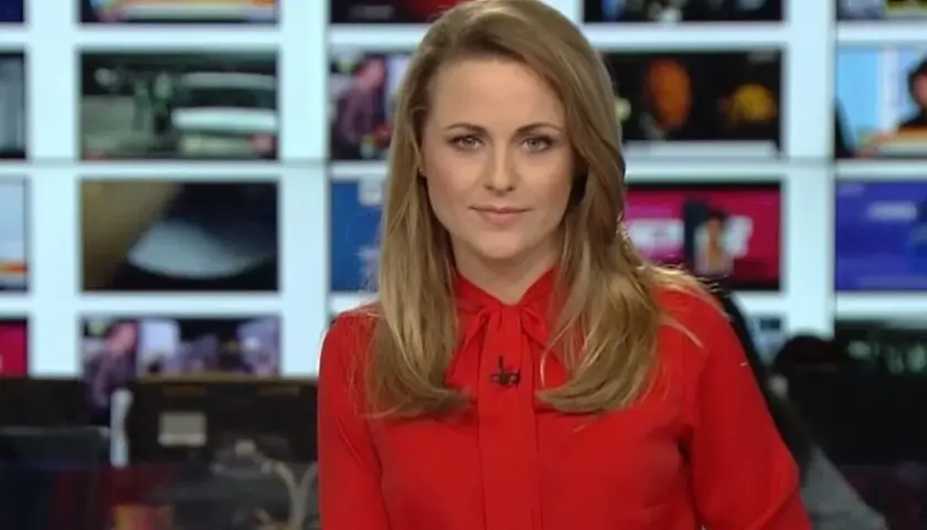 Marta Kłos, której wiek nie jest widoczny, w czerwonej bluzce z kokardą, na tle ekranów informacyjnych.