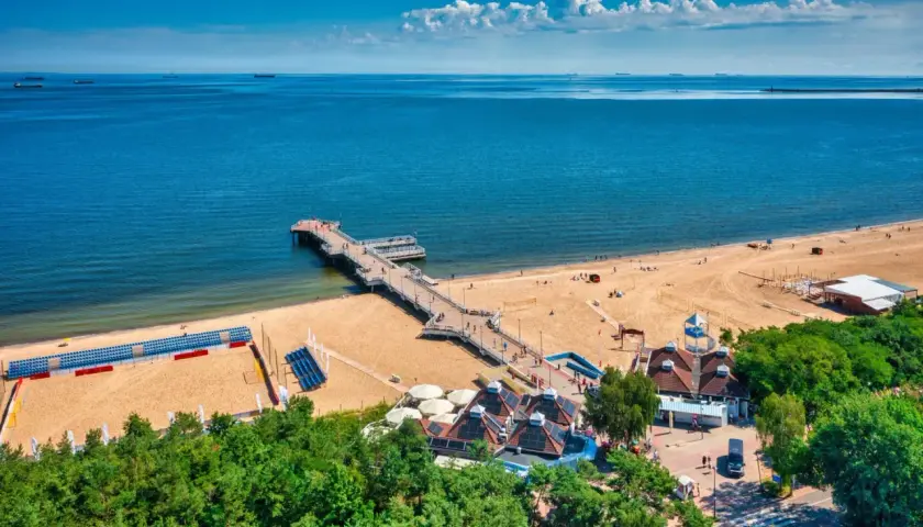 Piękny dzień na plaży w Polsce. Molo wchodzi w głąb morza, a piasek jest pełen ludzi cieszących się wakacjami.