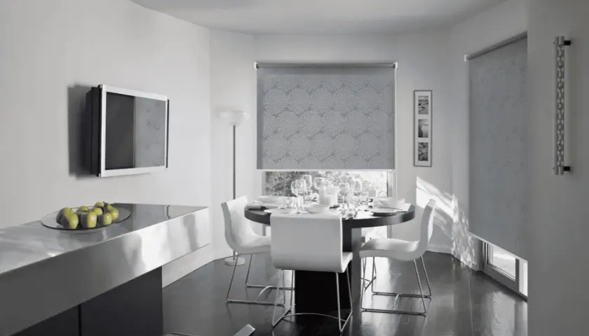 Tende antirumore funzionano: un elegante soggiorno con tavolo da pranzo apparecchiato e tende a rullo con motivo floreale che bloccano il rumore esterno.
