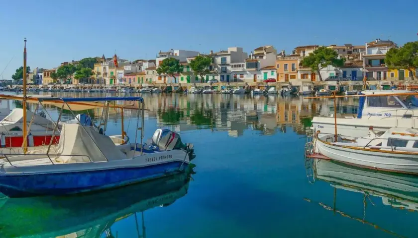 Boote im Hafen von Portocolom, Mallorca, spiegeln sich im ruhigen, blauen Wasser vor bunten Häusern.