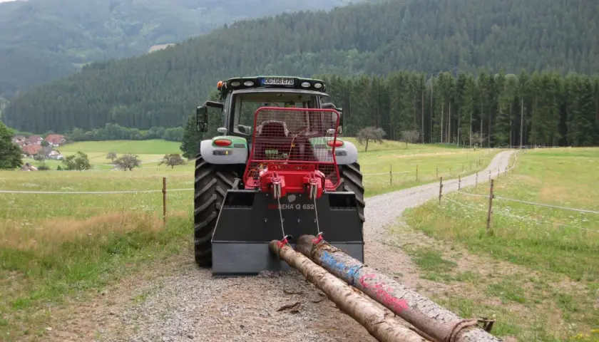 Traktor mit angehängter seilwinde forst zieht zwei Baumstämme über einen Schotterweg, umgeben von grünen Wiesen und einem dichten Wald.