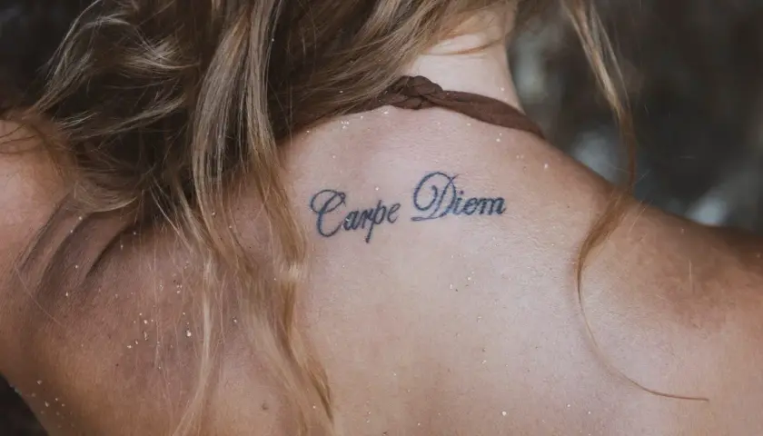 Tatuaże z napisami na karku: "Carpe Diem" na skórze, otoczone jasnymi włosami i drobnym piaskiem.