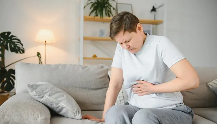 Kobieta siedzi na kanapie, trzymając się za brzuch. Jej twarz wyraża ból, co może sugerować przewlekłe zapalenie trzustki.