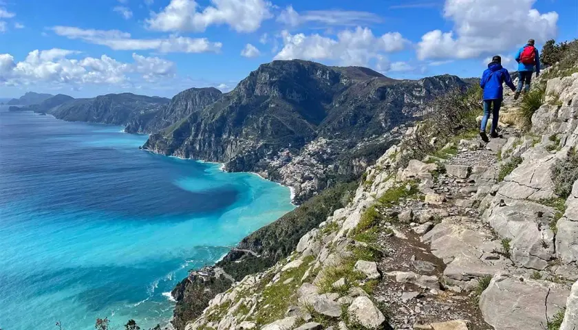 Hikers on the "walk of the gods" trail, with stunning turquoise waters and coastal villages below.
