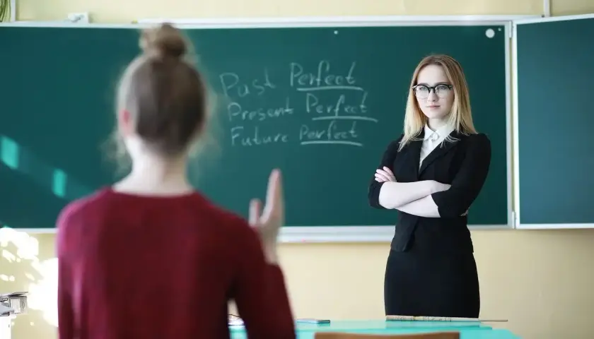 Nauczycielka w okularach stoi przed tablicą z zapisanymi czasami gramatycznymi.