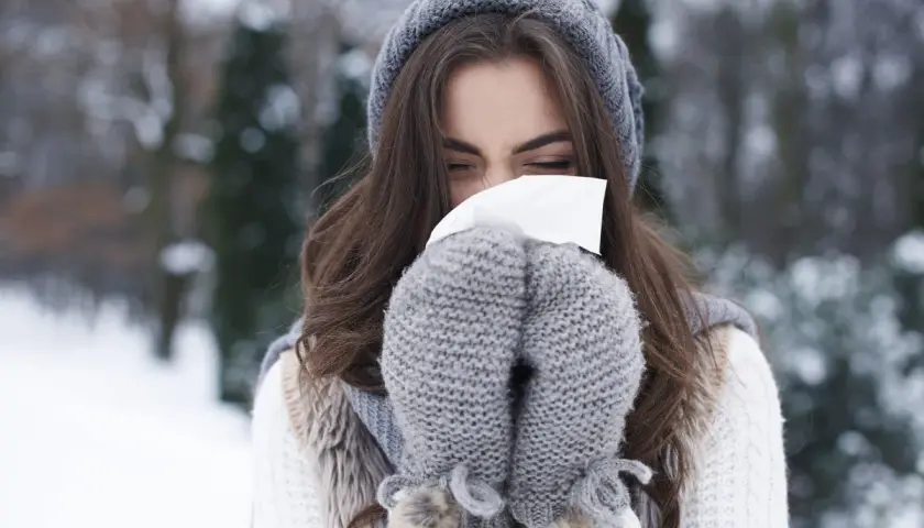 Femme en bonnet et moufles d'hiver se mouchant, un rappel pour prévenir le rhume.