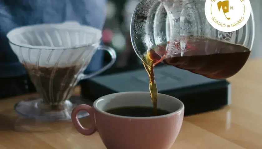Frisch gebrühter Kaffee fließt aus einer Glaskaraffe in eine rosa Tasse. Ein V60-Filter mit Kaffee steht bereit. Das perfekte Kaffee Rezept für Genießer.