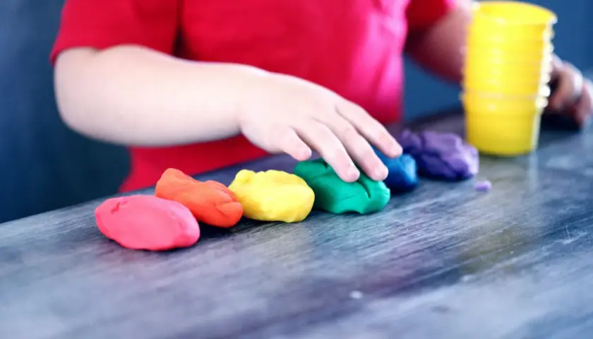 Dziecko w czerwonej koszulce bawi się plasteliną w tęczowych kolorach. To idealny obrazek do materiałów o kursie na opiekuna w żłobku.
