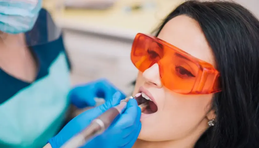 Pacjentka w okularach ochronnych poddaje się zabiegowi stomatologicznemu. Dentysta w rękawiczkach używa wiertła. Nowoczesne znieczulenie komputerowe zapewnia komfort.
