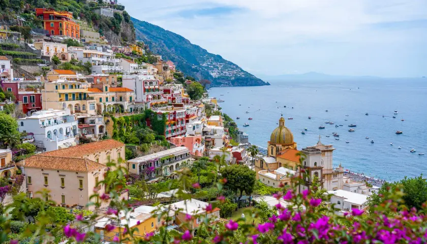 Colorful buildings cascade down a cliffside along the Amalfi road, overlooking a sparkling blue sea dotted with boats.