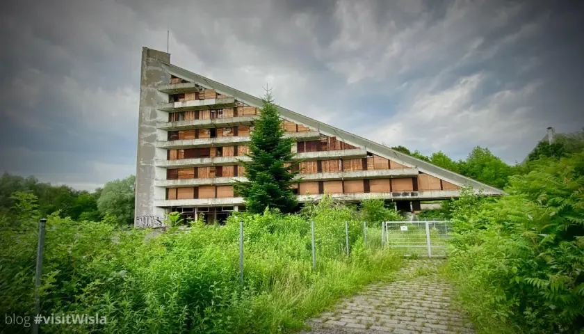 Opuszczony budynek o trójkątnym kształcie, z oknami zabitymi deskami, otoczony bujną zielenią. Idealne urbex miejsca dla poszukiwaczy przygód.