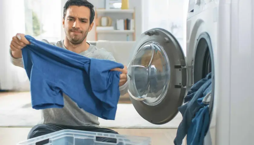 Hombre mira con preocupación una camiseta azul, parece que la viscosa encoge. La lavadora está abierta con ropa dentro.