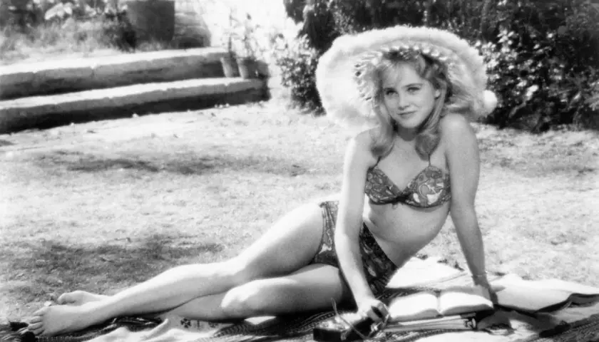 Młoda kobieta w bikini i kapeluszu leży na trawie, czytając książkę. Scena jak z letnich filmów erotycznych.