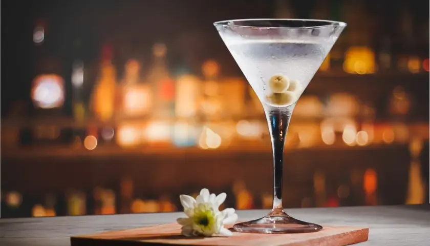 Martini extra dry z oliwkami, idealny do wieczornego relaksu. Na blacie stoi kieliszek z drinkiem i biały kwiatek.