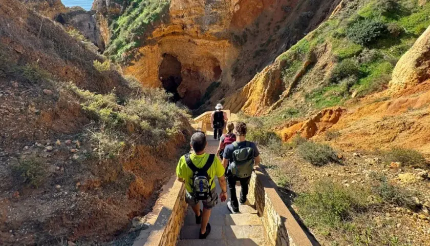 Hikers descend a stone staircase on a scenic trail in Portugal, leading towards a large cave entrance nestled within rugged, sun-drenched cliffs.